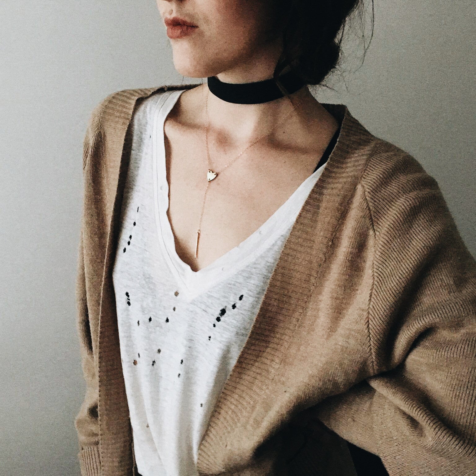 a close up of a woman wearing a ripped white tee shirt and brown cardigan with a thick black chocker necklace an a gold heart lariat necklace