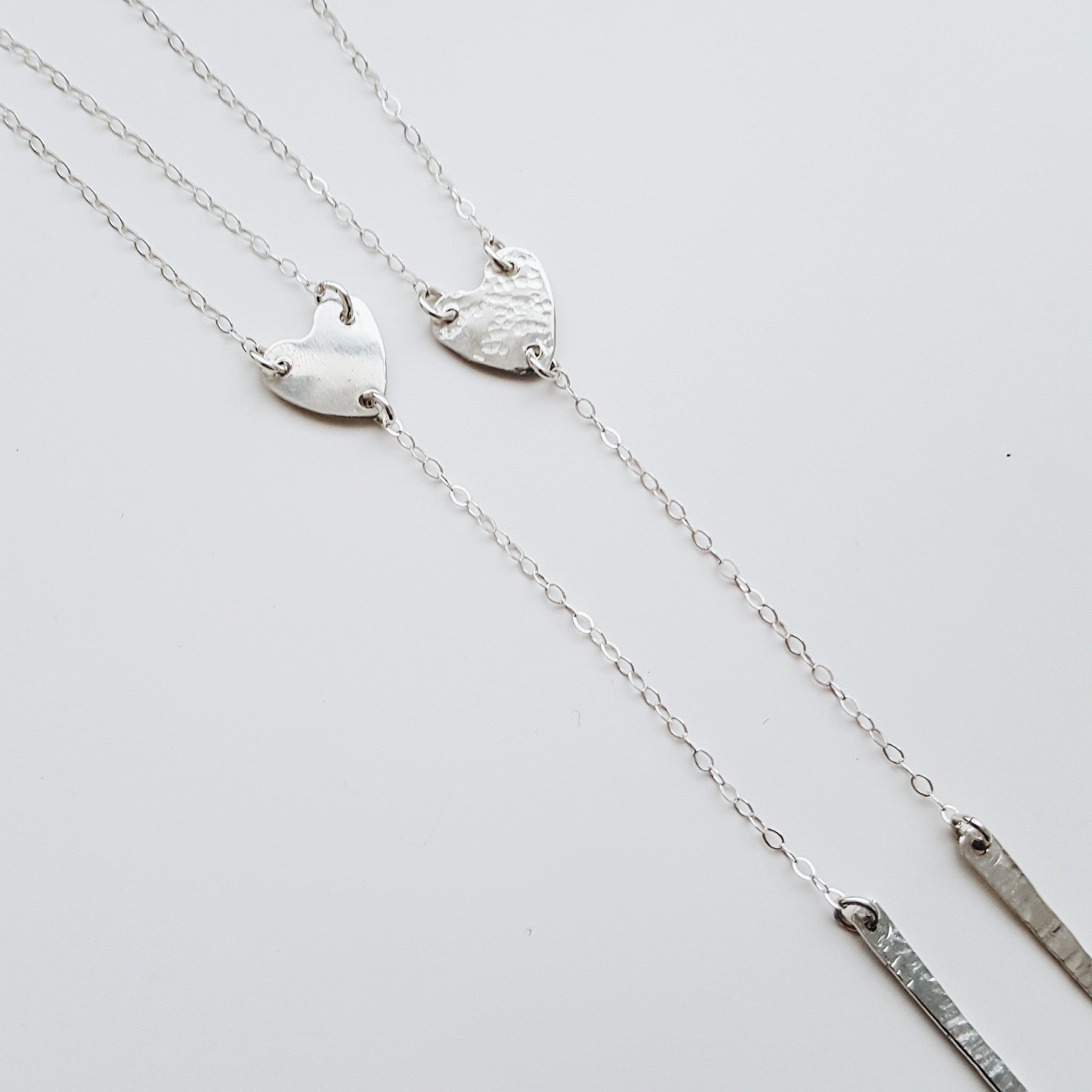 a silver heart lariat necklace with a silver chain and simple silver bar on a white background
