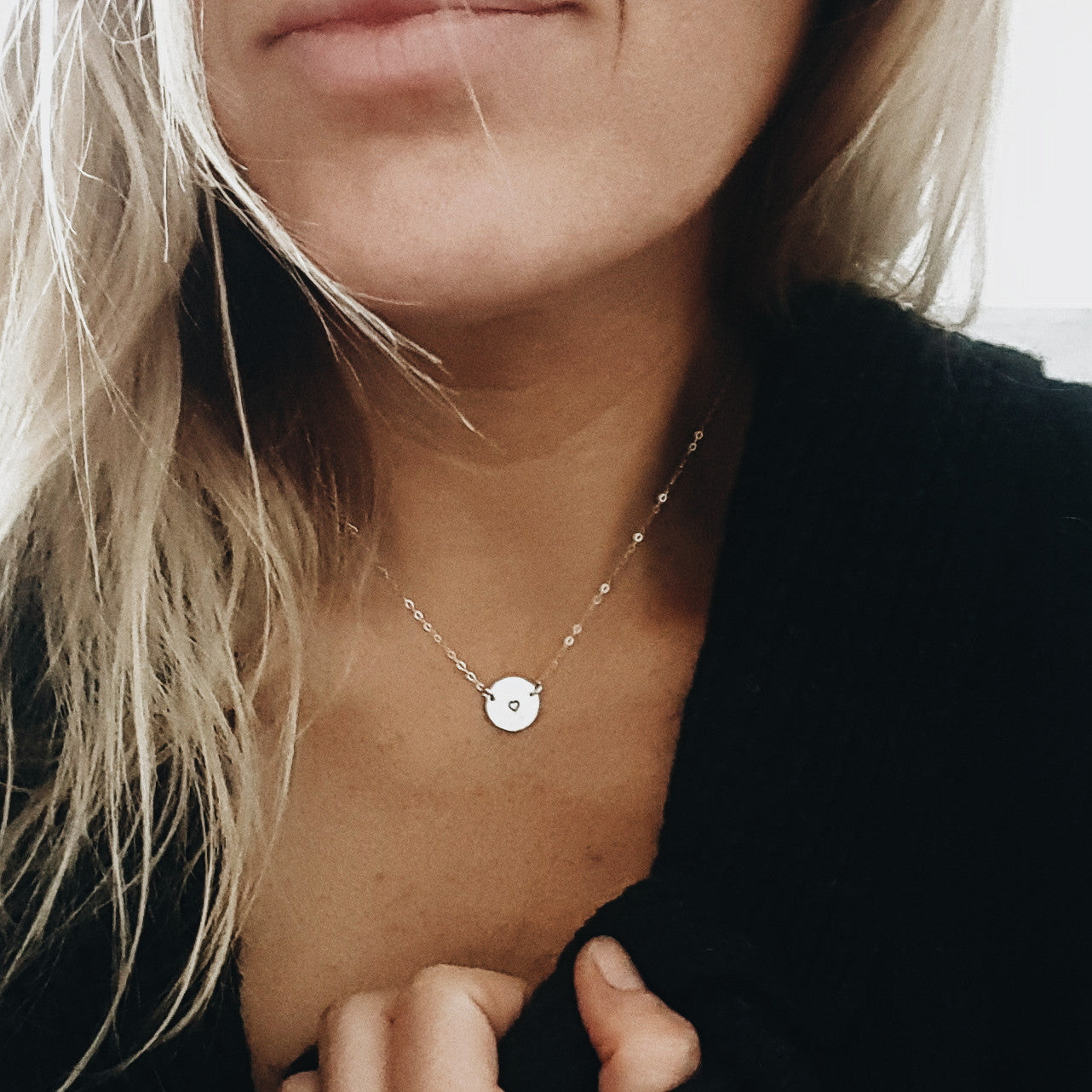 a close up of a woman wearing a black sweater and a small round gold pendant with a small heart stamped in the middle on a gold chain