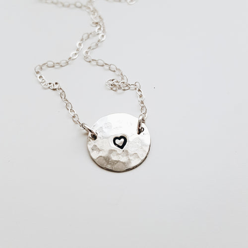 a round silver pendant with a small heart stamped on the centre and a silver chain shown on a white background