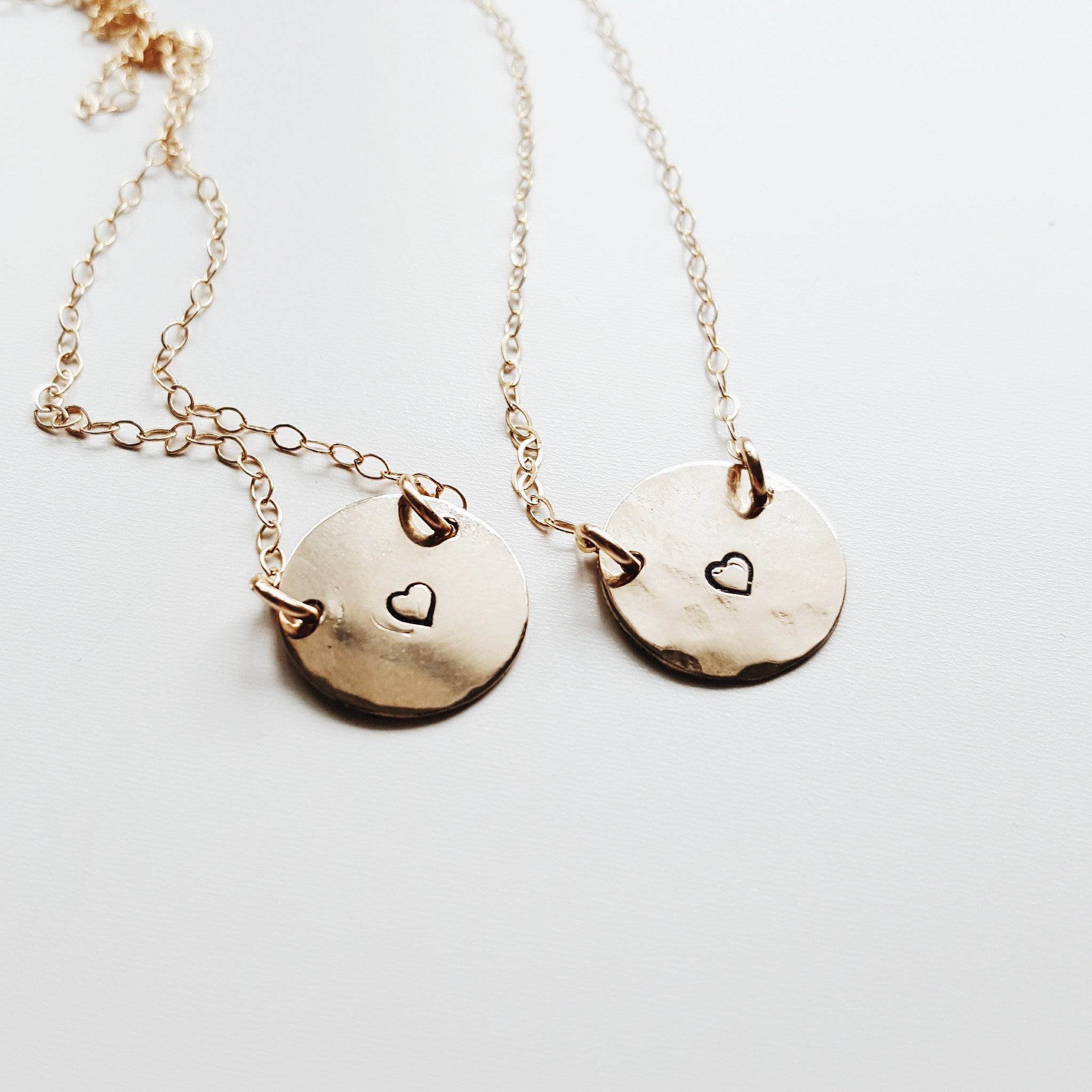 two small gold round pendants with a small heart stamped on the middle of each, on gold chains shown on a white background