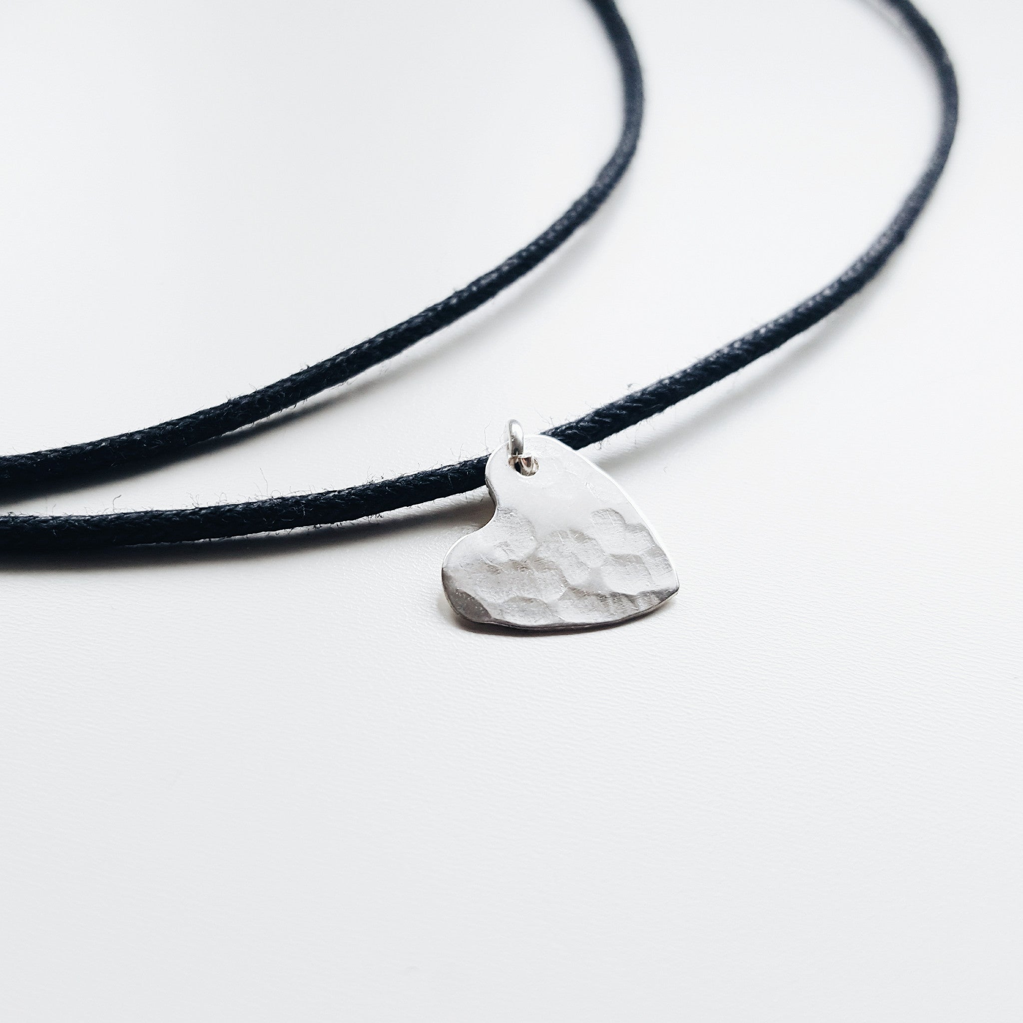 a black rope necklace with a small silver hammered heart necklace on a white background