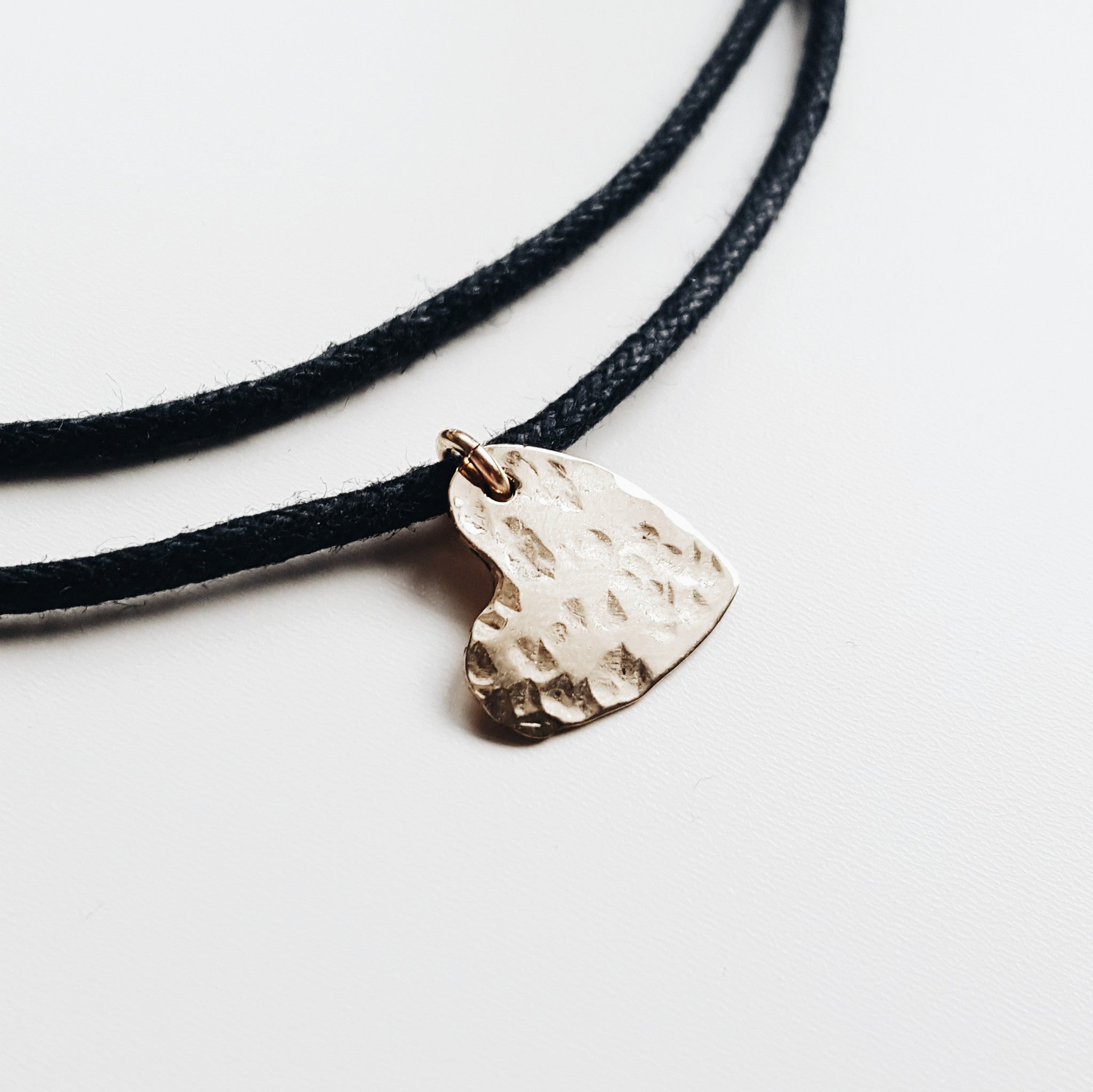 a black rope necklace with a small gold hammered heart necklace on a white background