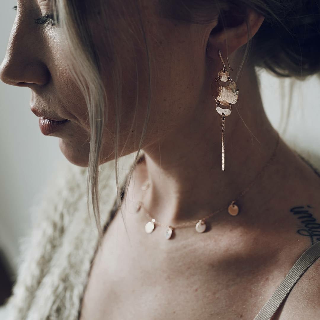 a side profile of a woman wearing a beige knit cardigan and gold hammered metal earrings, with a gold necklace of dangling metal circles