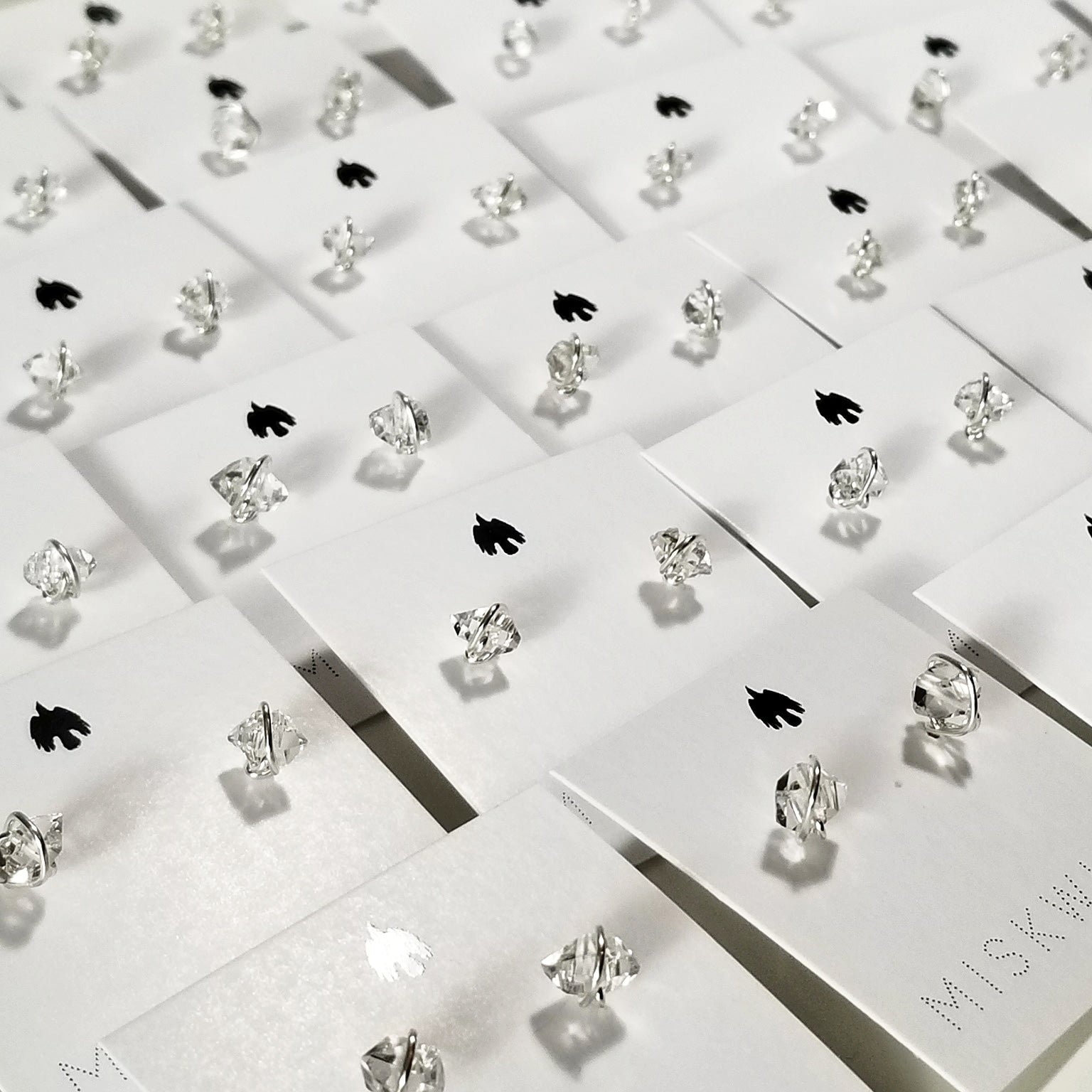 a group of silver herkimer diamond stud earrings on white cardstock holders with a small black raven logo