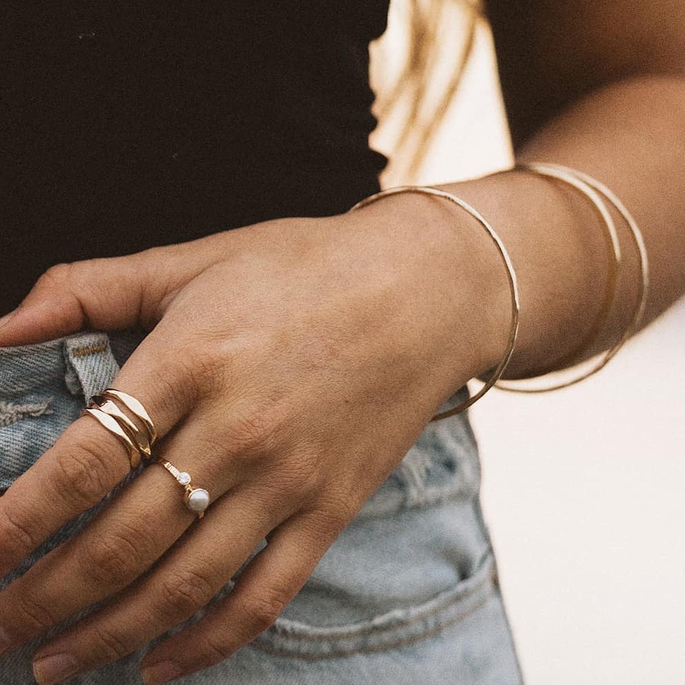 a close up photo of a hand posed on blue jeans wearing 3 stacked gold rings, a small gold pearl ring and three gold stacked bracelets 