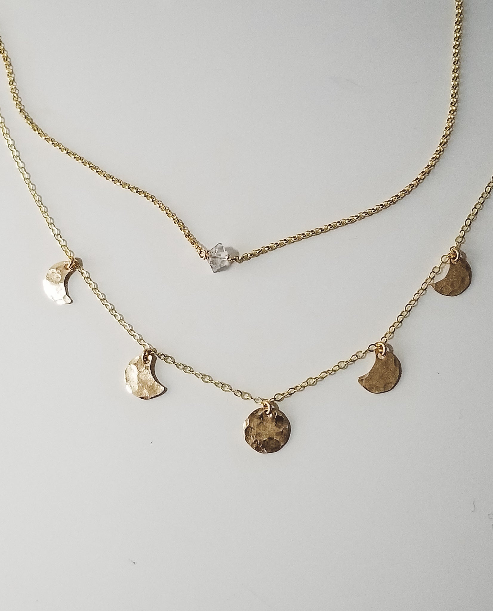 a herkimer diamond on a gold chain and a hammered metal moon phase necklace in gold on a white background