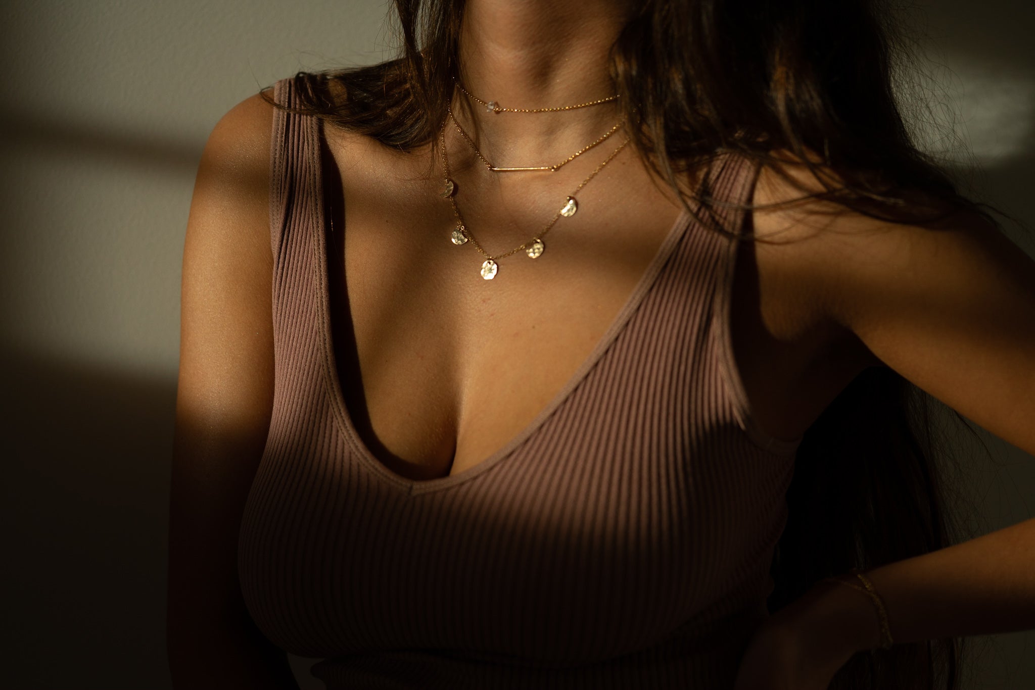 a close up of a woman posing in a dark beige tank top with layered gold necklaces in the sun