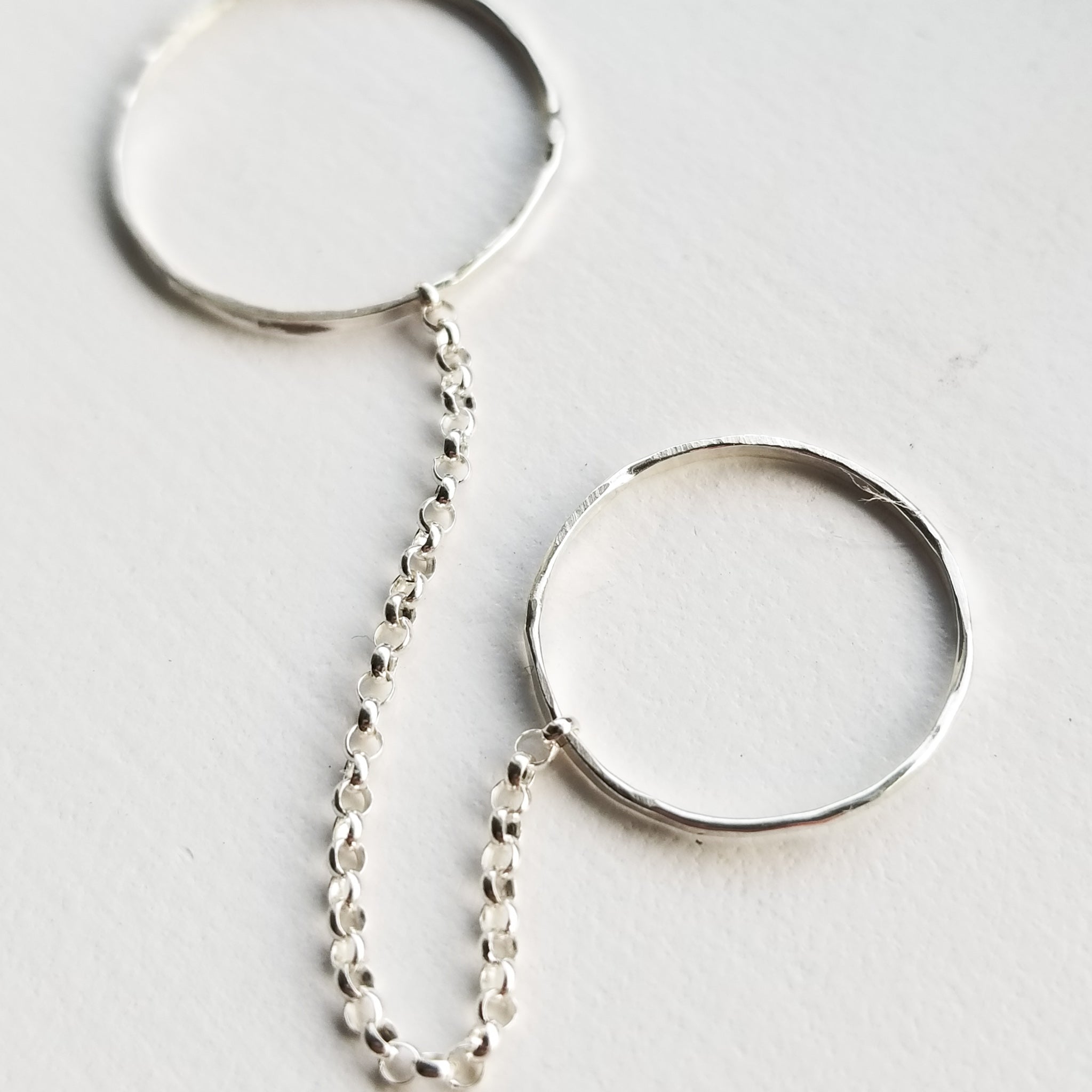 a set of two silver band rings connected by a silver chain on a white background
