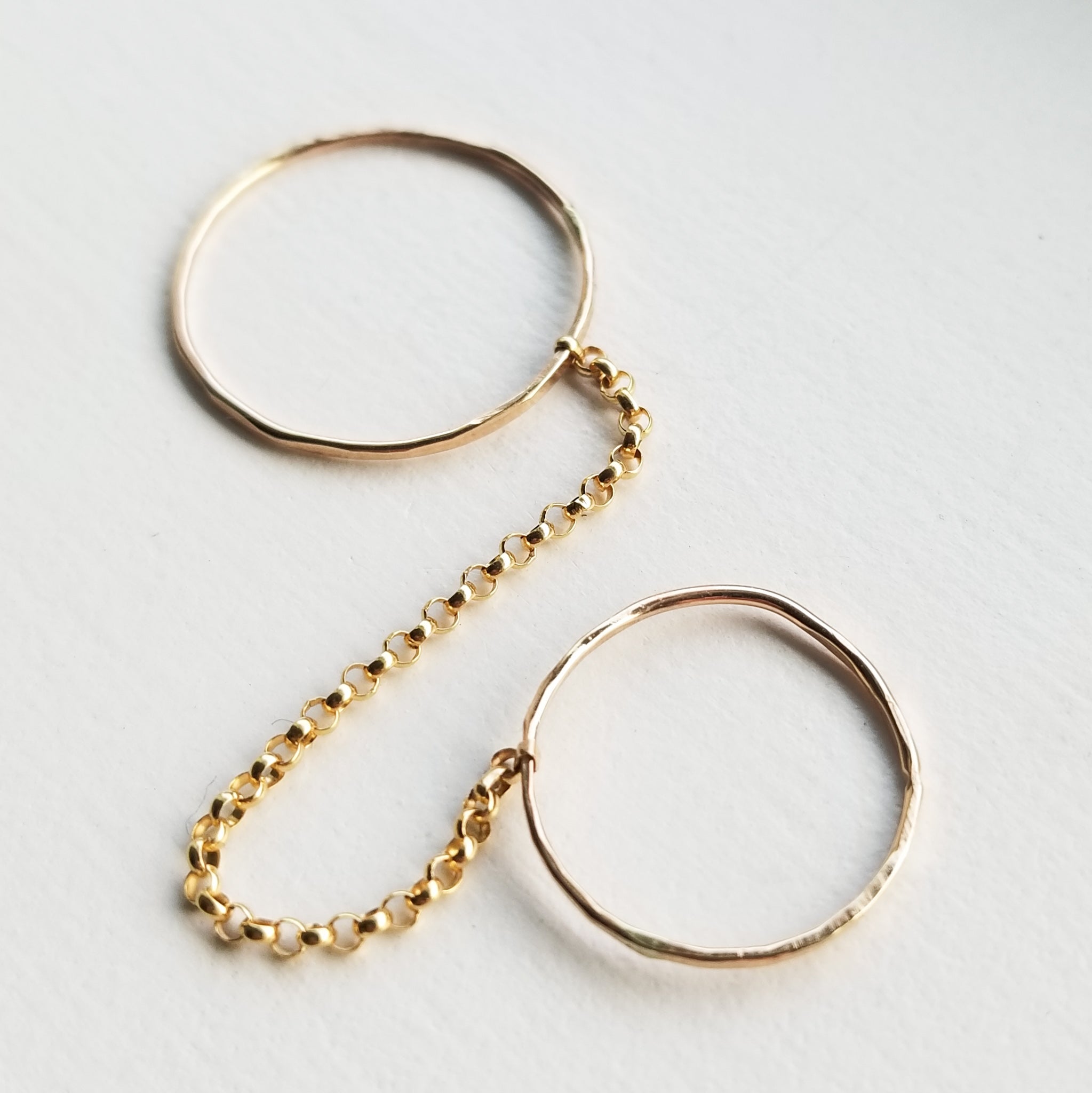a set of two gold simple band rings connected by a gold chain on a white background
