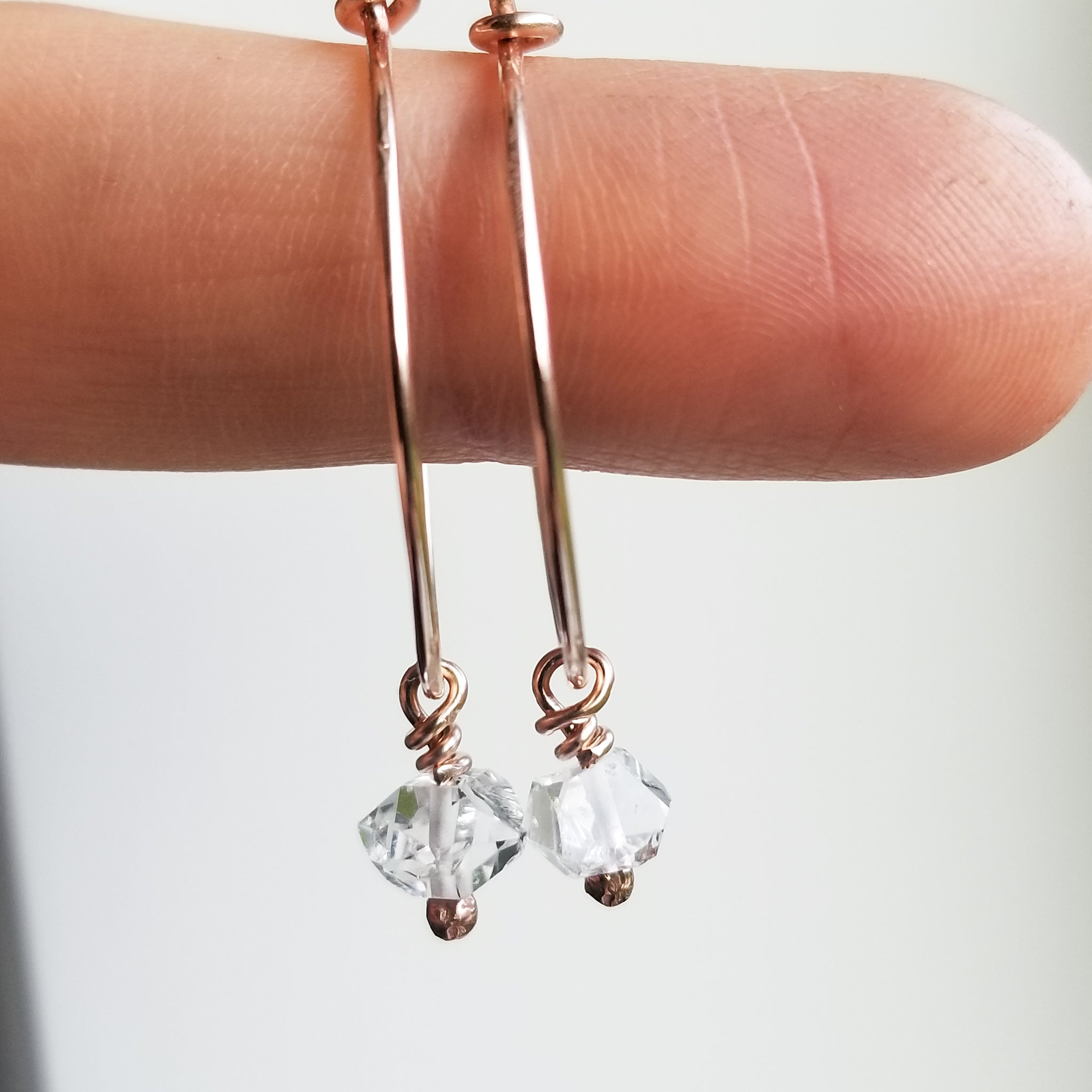 a finger holding small rose gold hoops with dangly hermiker diamonds on a white background