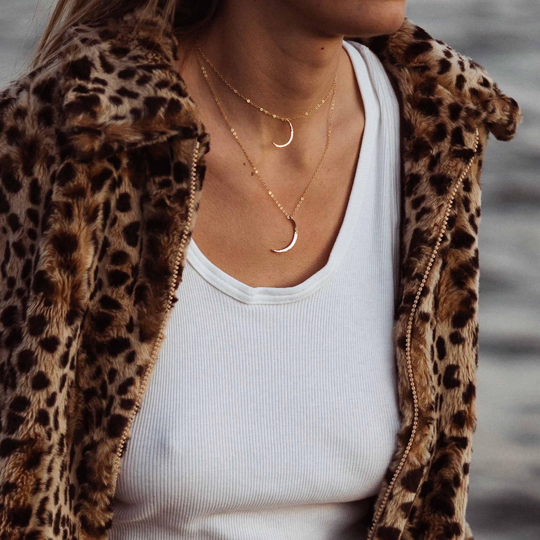 a close up of a woman wearing two gold crescent moon shaped pendant necklaces on gold chains and a white tank top with a leopard print jacket