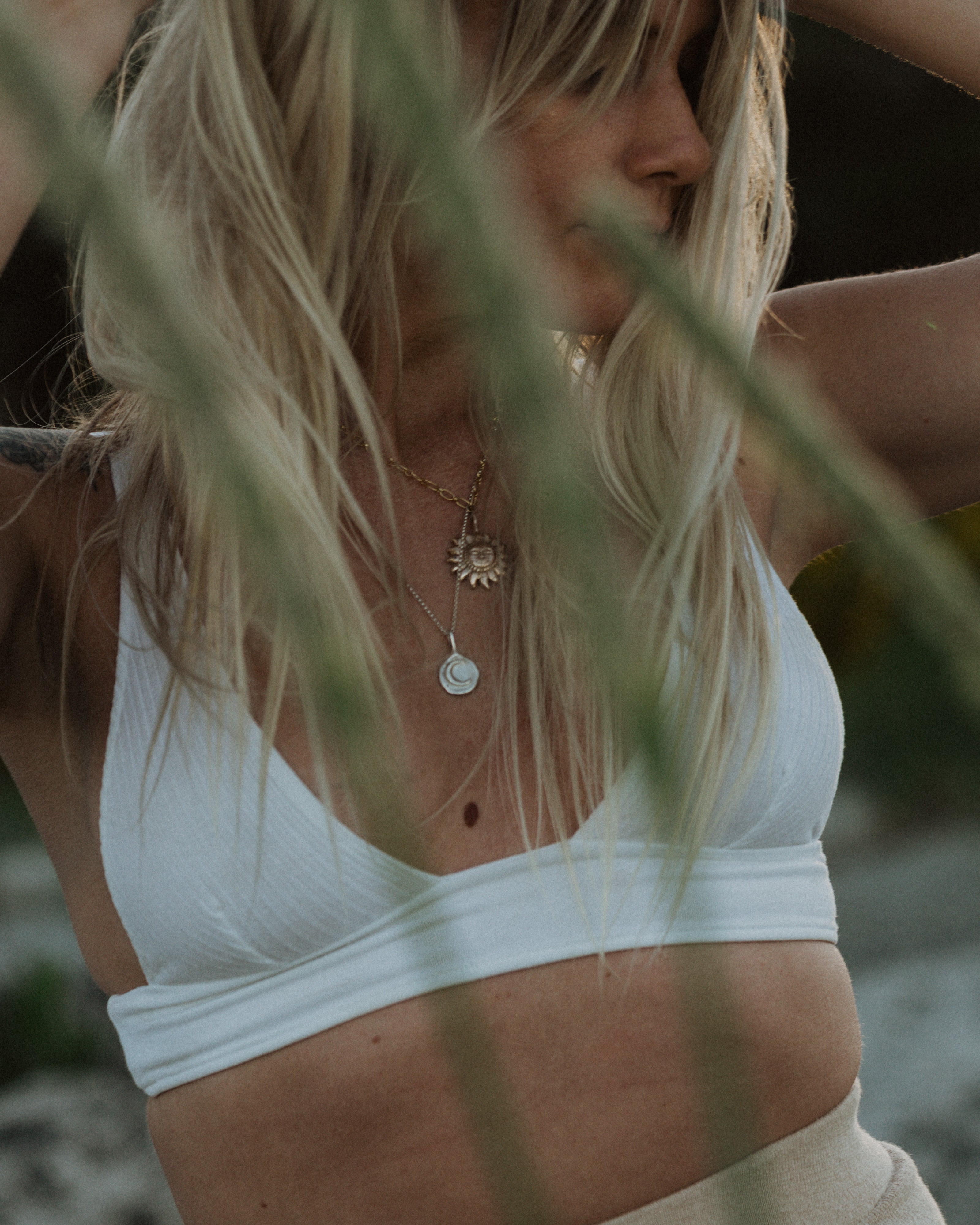 a woman wearing a white top in the sun wearing a gold sun pendant necklace and silver moon pendant necklace, peaking through green leaves