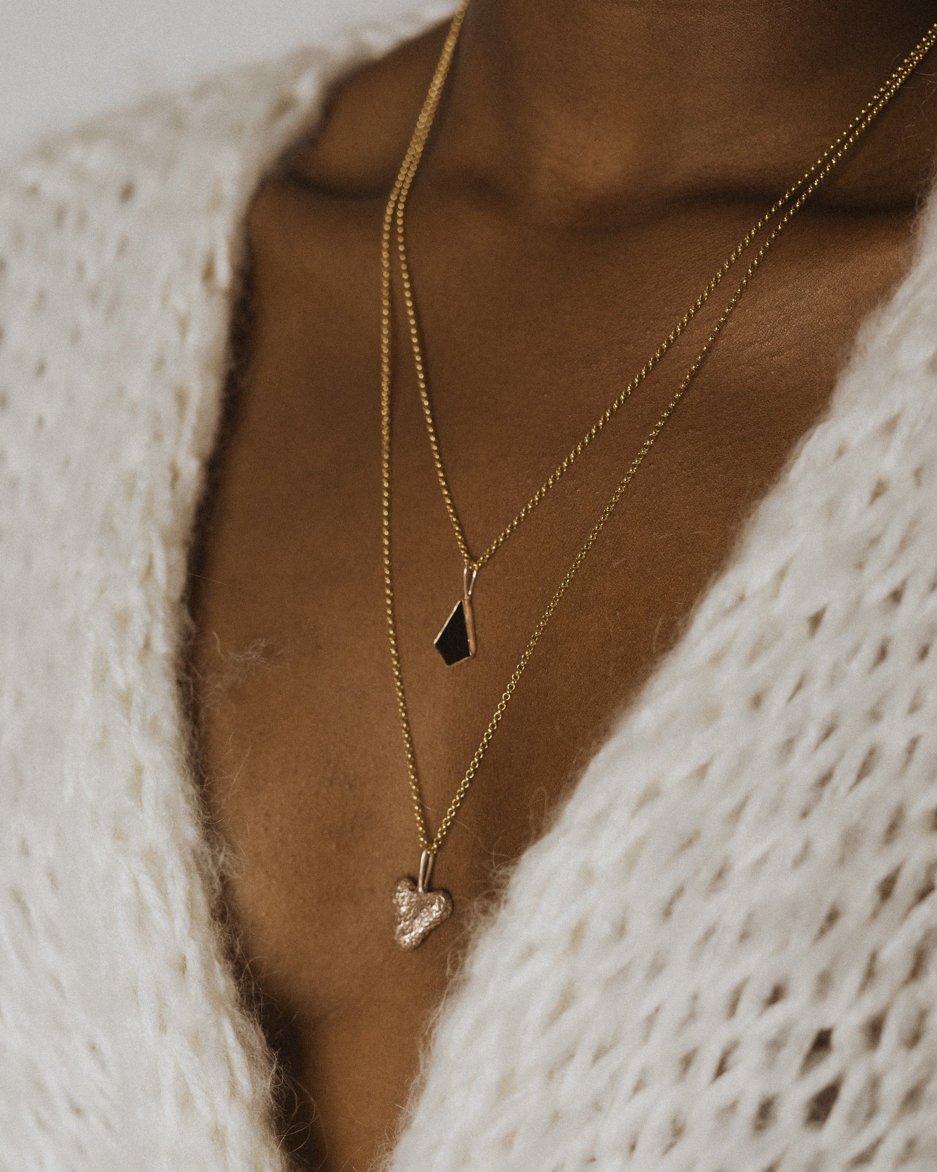 close up of a womans neck wearing a small bronze coral heart pendant and a black stone pendant with a white knit cardigan