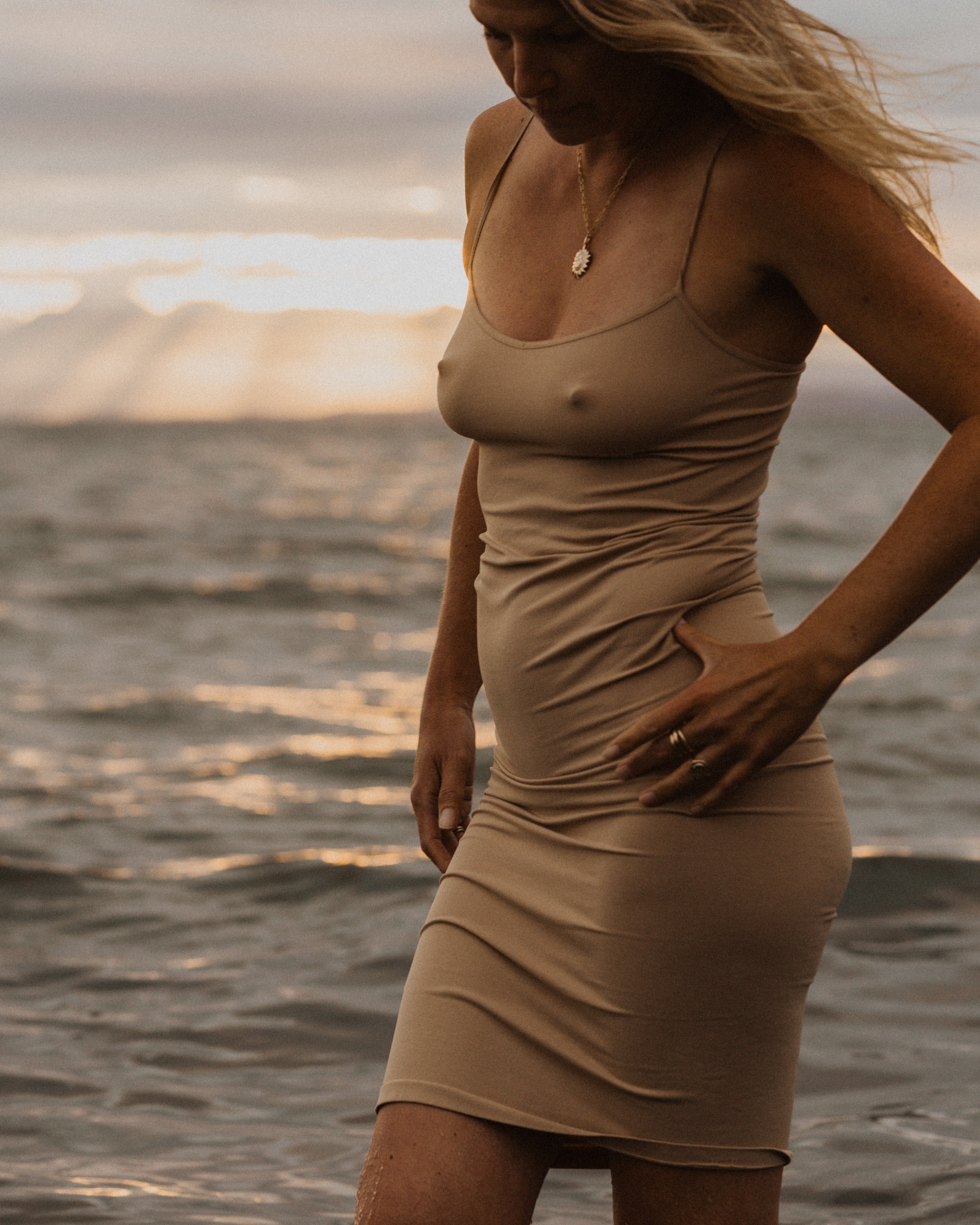 a woman standing in front of the ocean wearing a beige dress, a bronze sun pendant on a gold chain and stacked bronze rings on her fingers