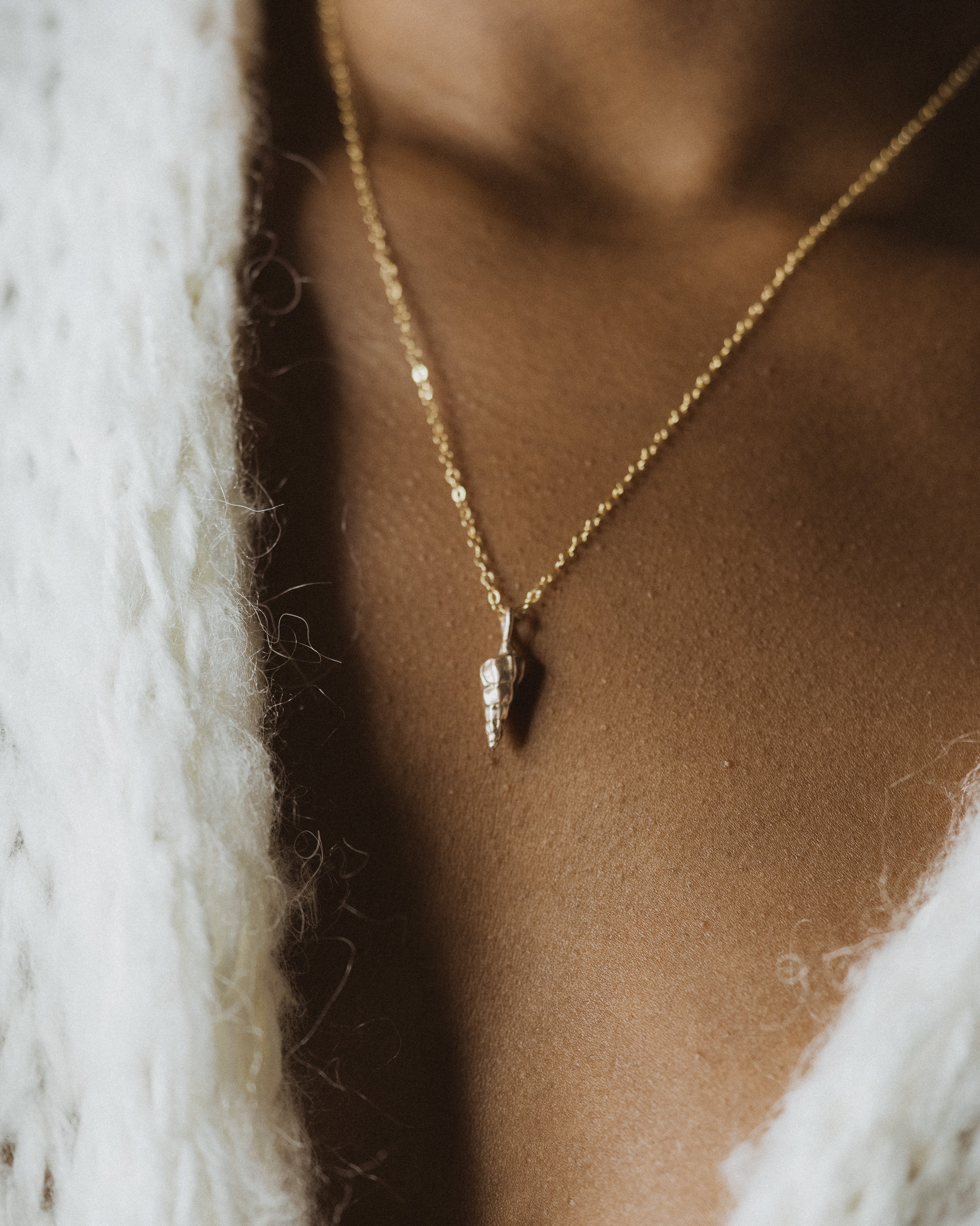 a close up of a woman's neck wearing a white knit cardigan and small bronze sea shell pendant necklace