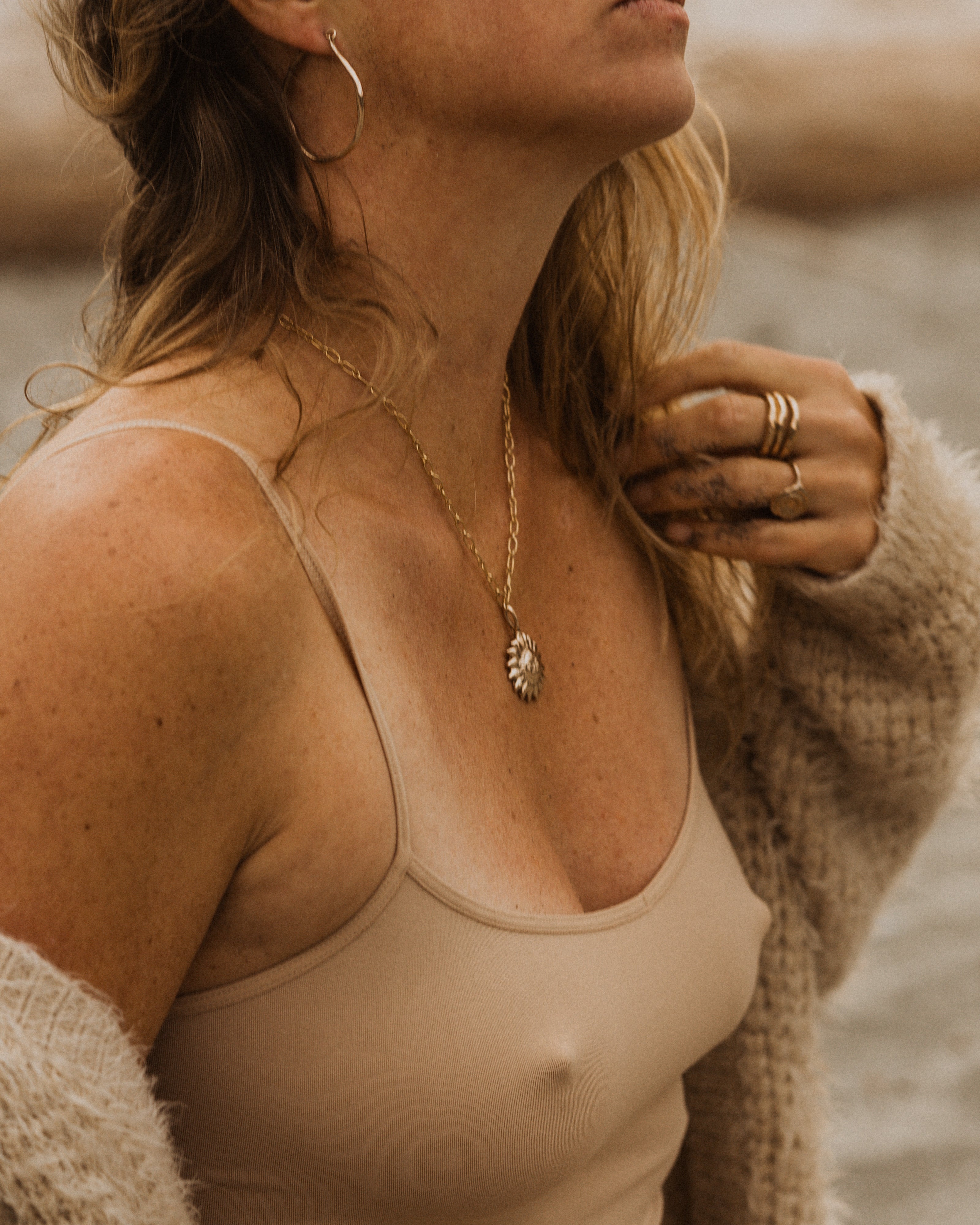 a side profile of a woman at the beach, wearing a beige tank top and beige knit sweater. She has a bronze sun pendant on a gold chain necklace, stacked bronze rings and bronze hoop earrings