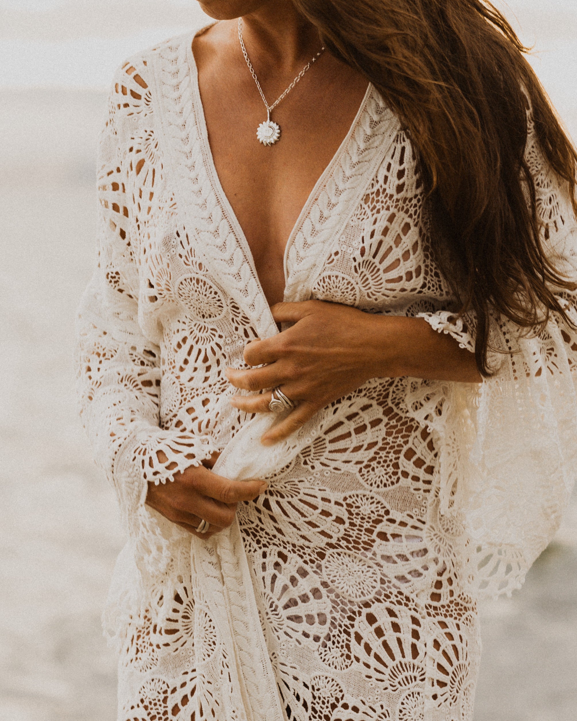 a woman at the beach, wearing a white lace rope and a silver sun pendant necklace, with layered silver rings on each hand