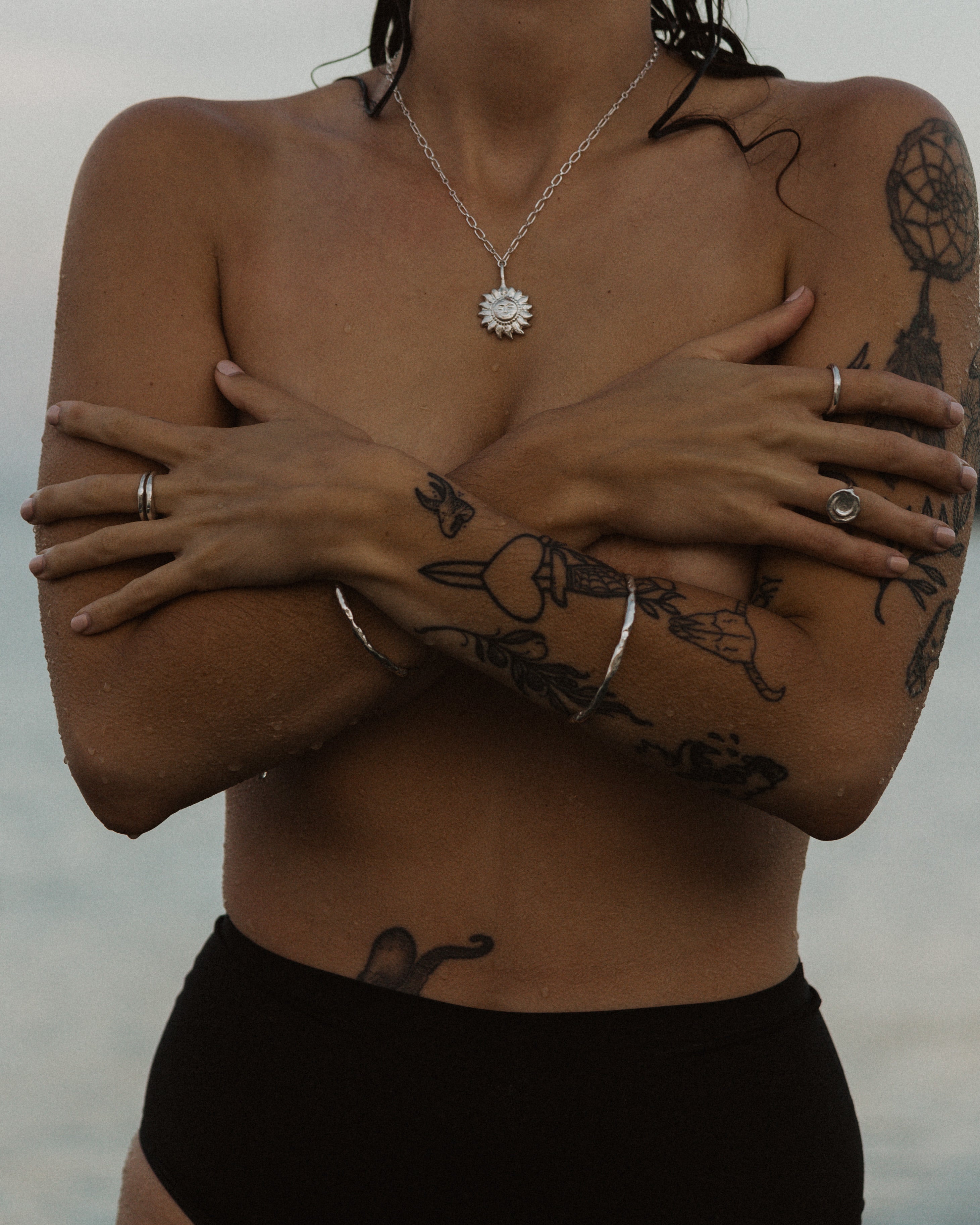 a cropped photo of a woman on the beach covering her chest, wearing stacks of silver jewelry and black bottoms