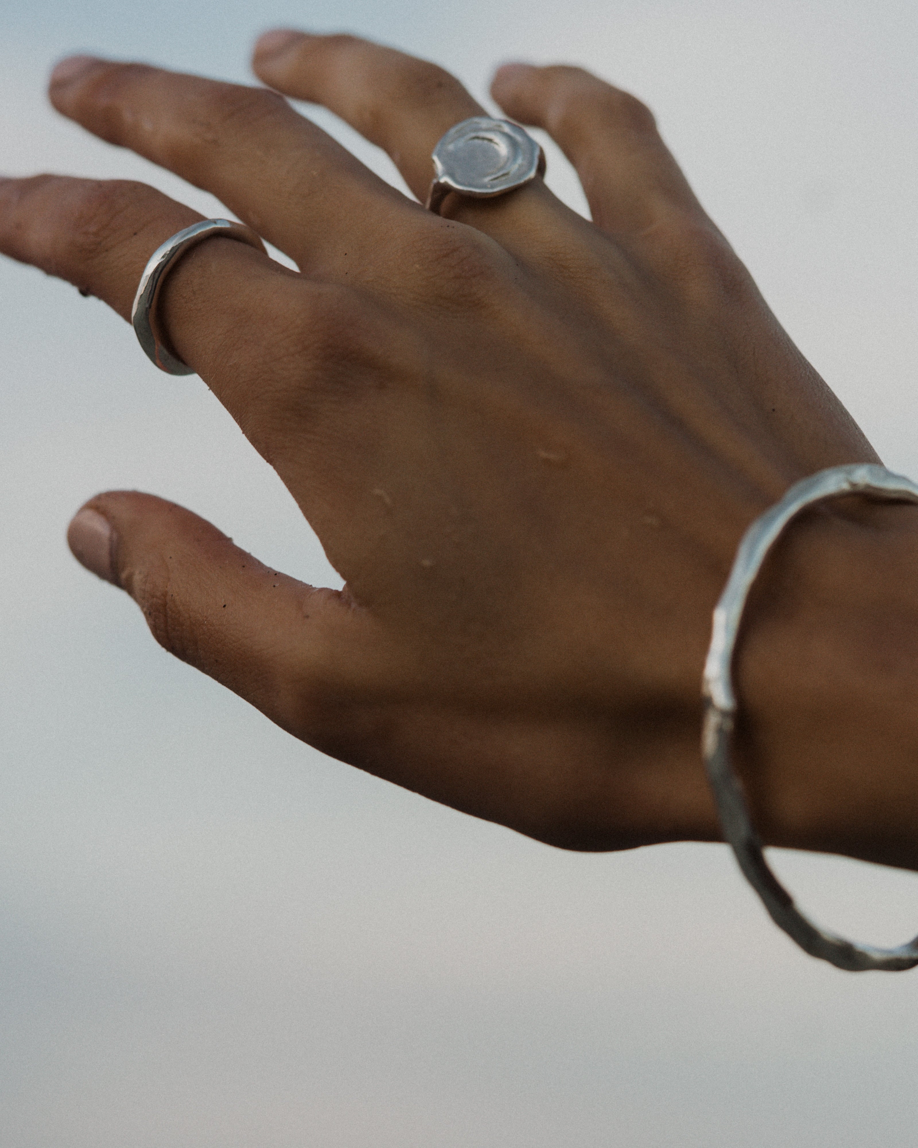 a close up of a hand held against the sky wearing silver hand formed rings and a silver bangle bracelet