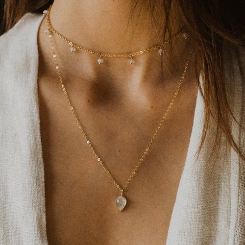 a close up of a woman's neck wearing a long gold chain with a tear drop opal pendant and a gold chocker with small beaded droplets