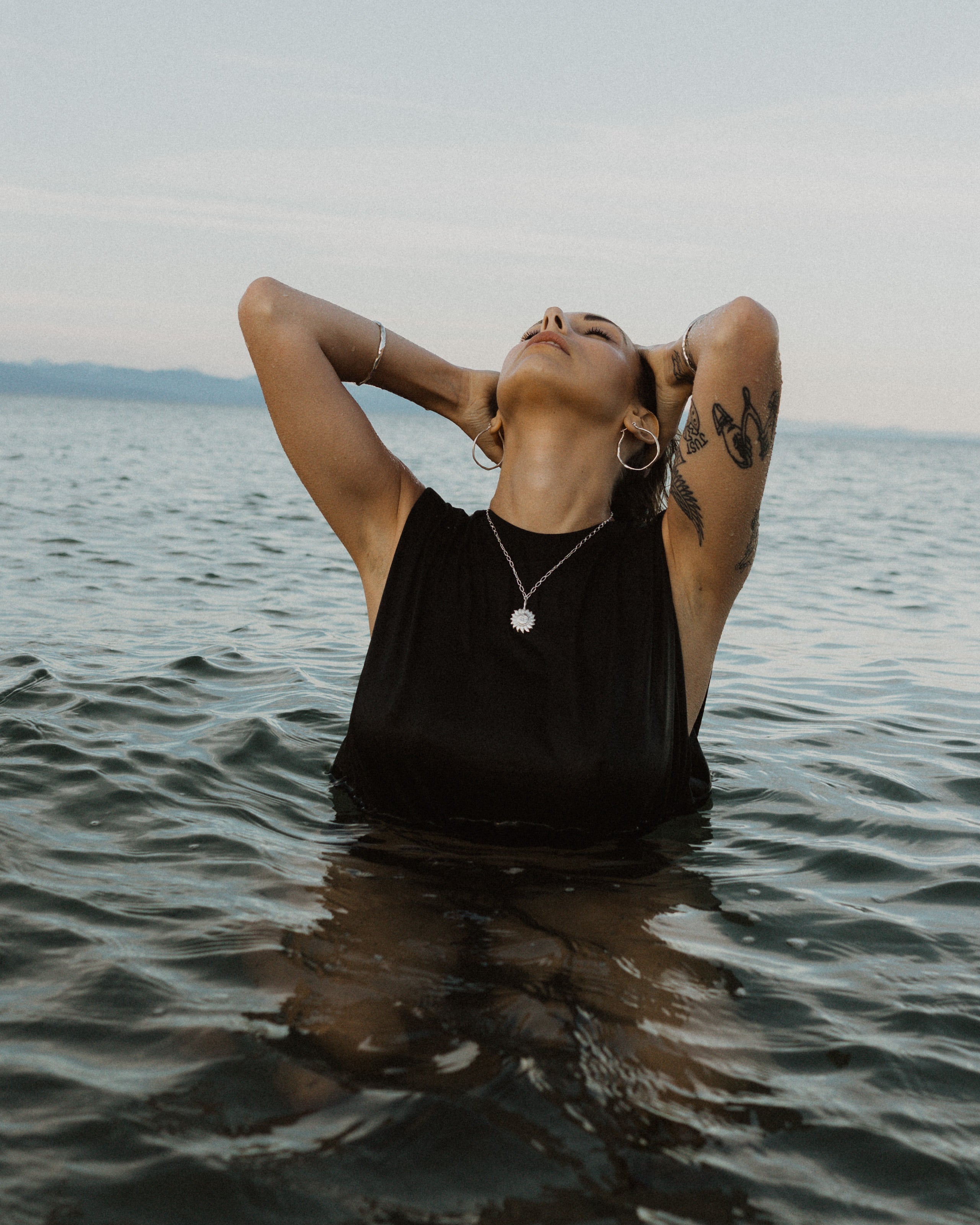 a woman kneeling in the water at the beach, wearing a black tank top and silver jewelry. Her jewelry includes a silver sun pendant necklace, silver hoop earrings and silver bangle bracelets