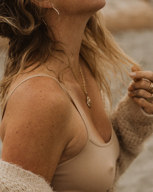 a close up of a woman wearing a beige tank top and beige knit sweater, with a bronze sun shaped pendant necklace, bronze hoop earrings and stacked bronze rings