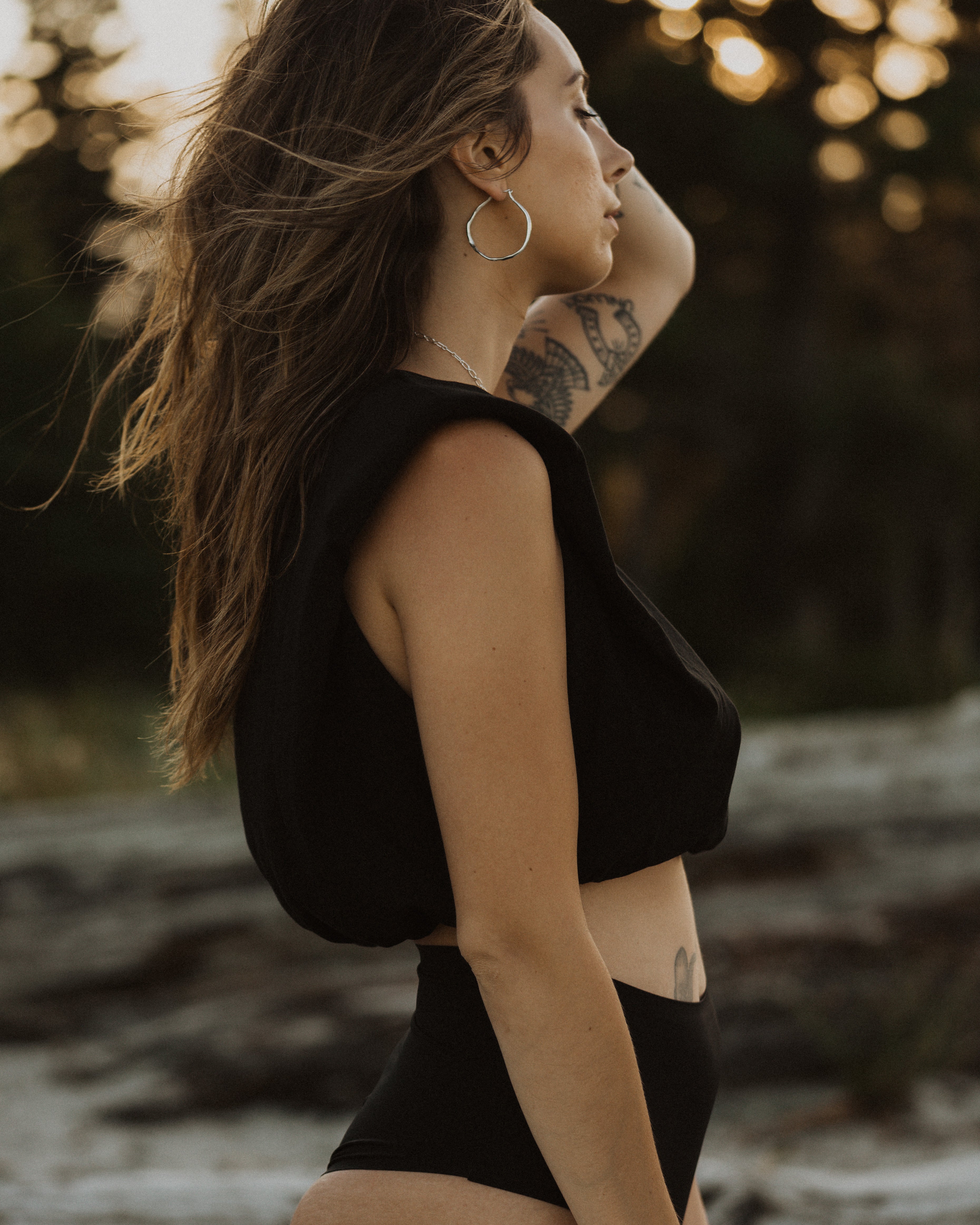 a side profile of a woman at the beach wearing a black top and black bottoms, with silver hoop earrings and a slight peak of a silver necklace