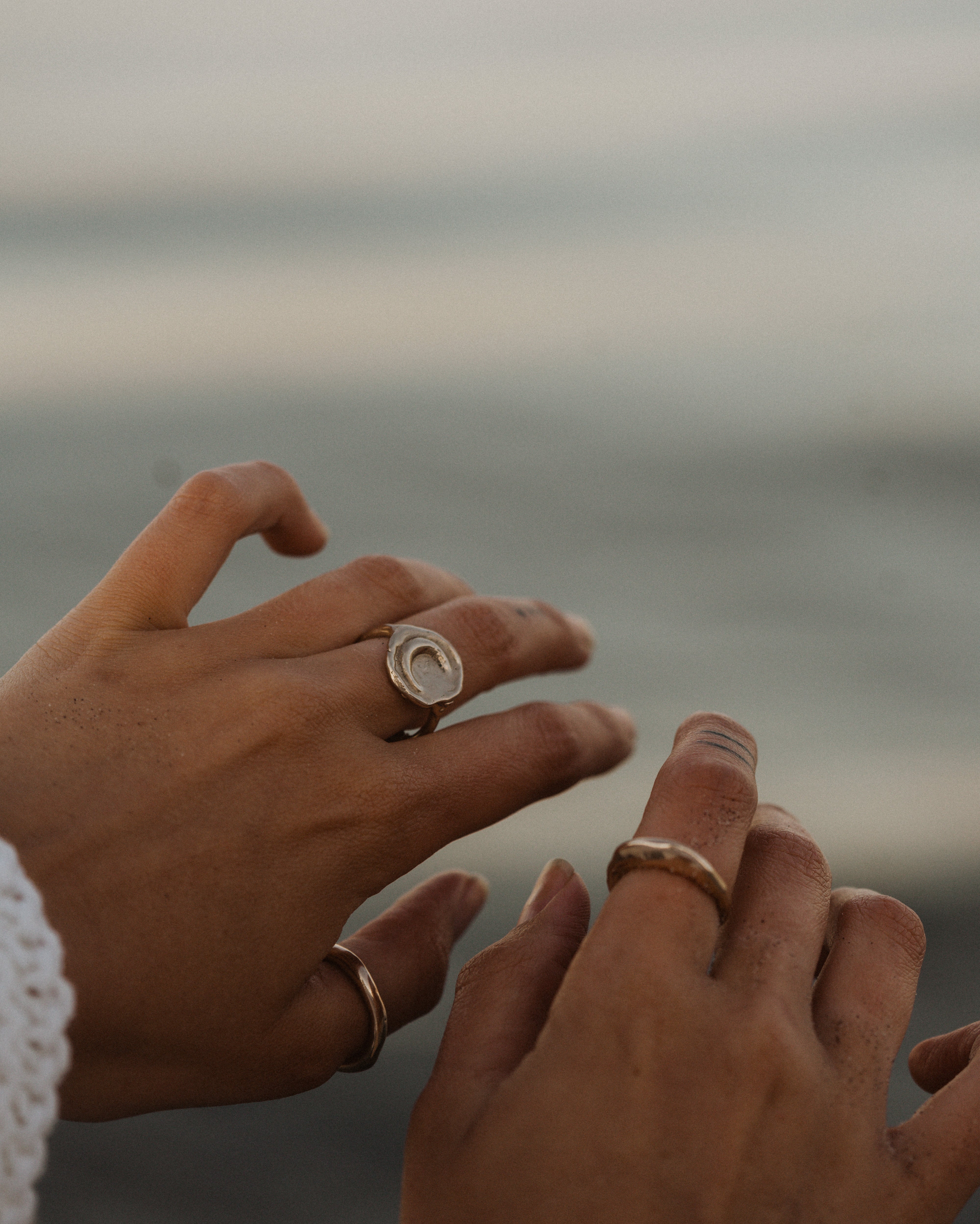two hands held in front of a blurred beach background wearing multiple bronze rings on each fingers
